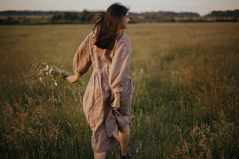 a woman carrying wild flowers and wearing a linen dress walking through a field