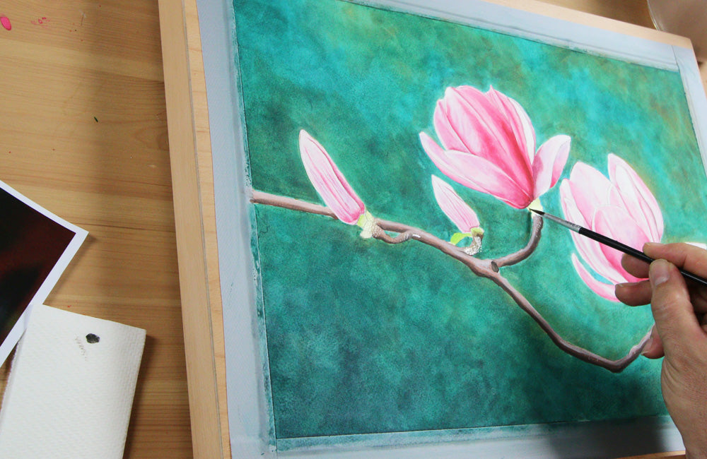 Hand painting pink watercolour flower with brush onto page taped to a wooden back board. 