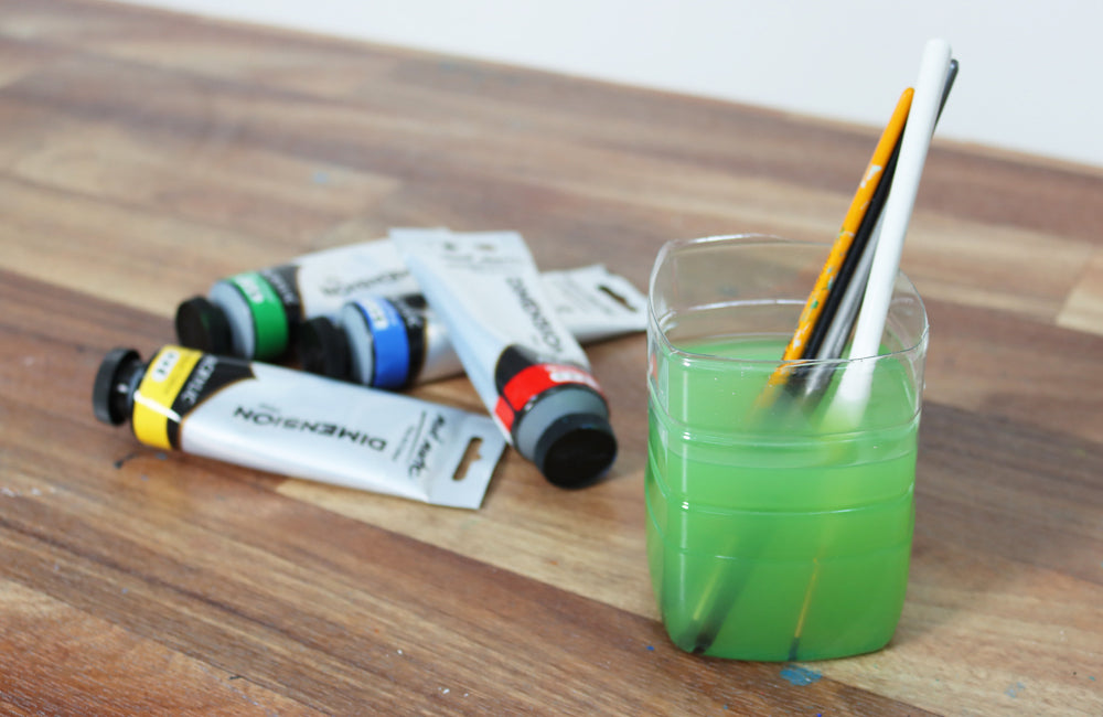 Yellow, green, blue and red paints on a wooden table next to a cup of green water and paint brushes.