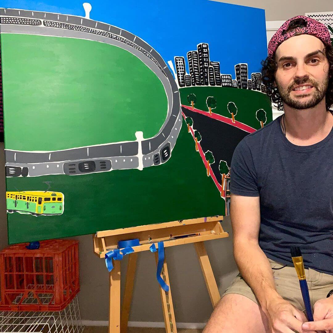 1. Artist Charles Nanopoulos holding a paint brush and sitting next to his abstract artwork of the Melbourne Cricket Ground.