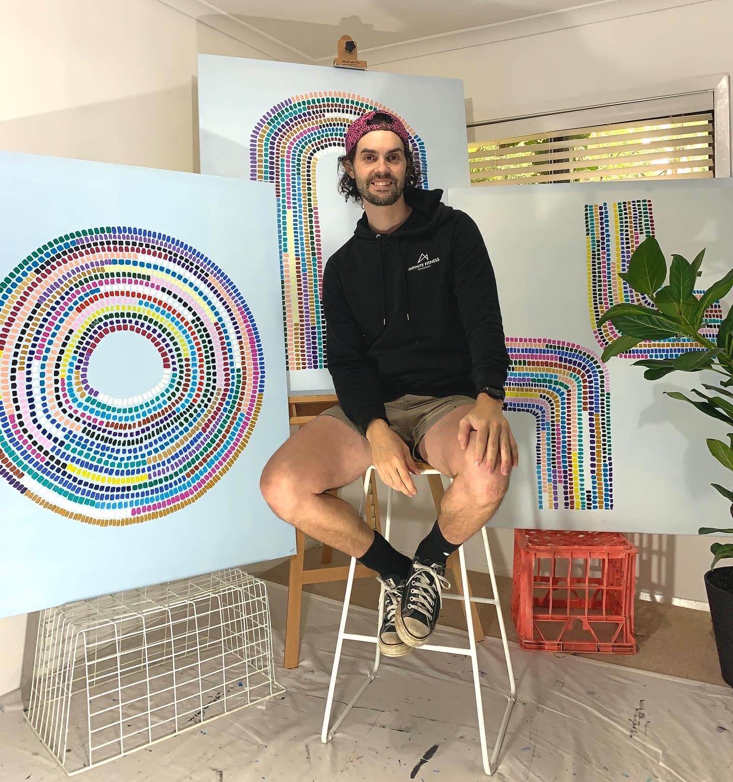 Artist Charles Nanopoulos sitting on a white stool in front of three large white geometric paintings on canvases.