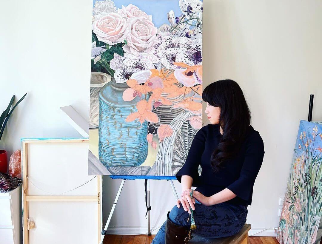 Artist Hsin Lin gazing happily at her artwork sitting on a canvas with an artwork of flowers in baskets painted on canvas