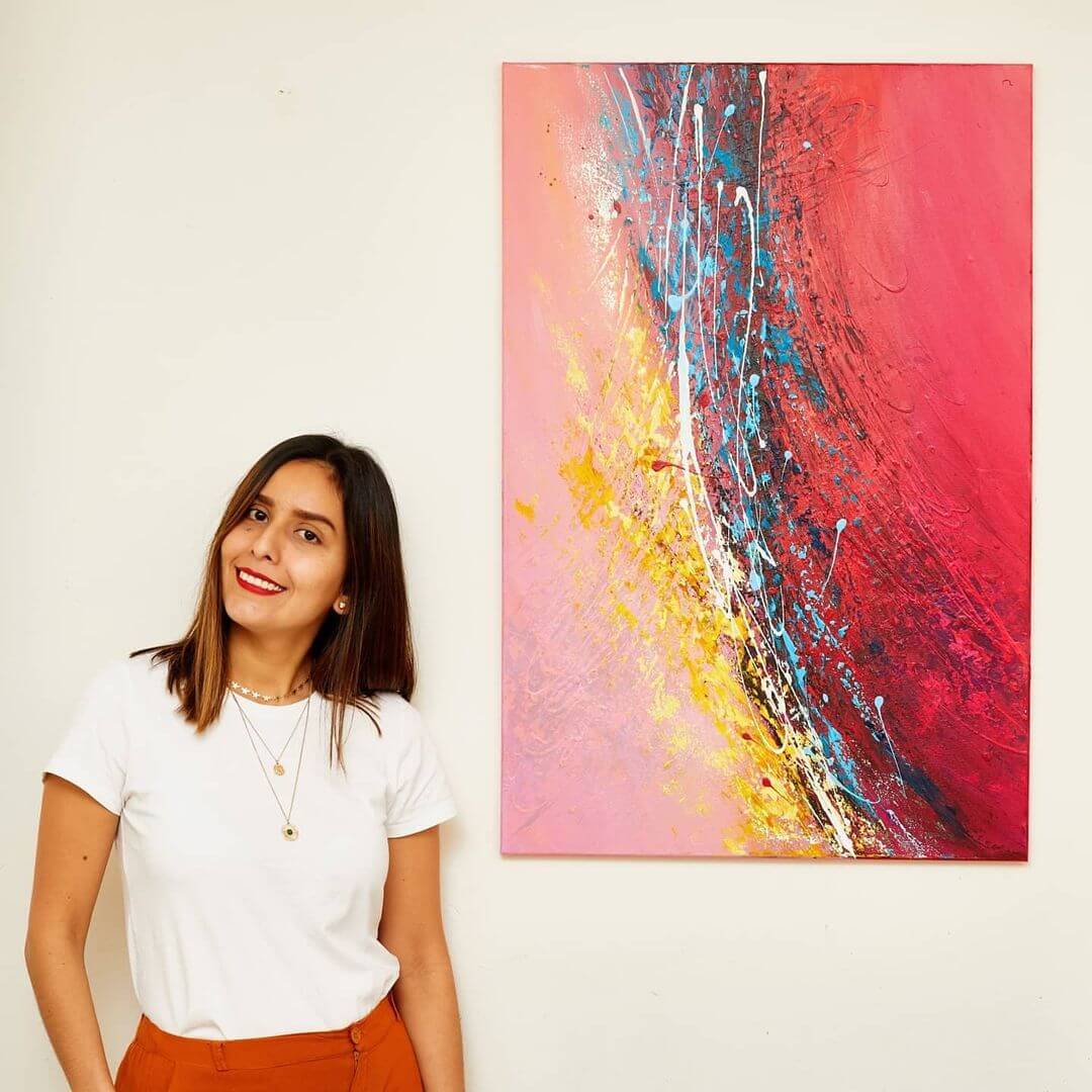 Artist Lina Mar standing and smiling next to a coloured canvas on a wall in a white t-shirt and gold jewellery.