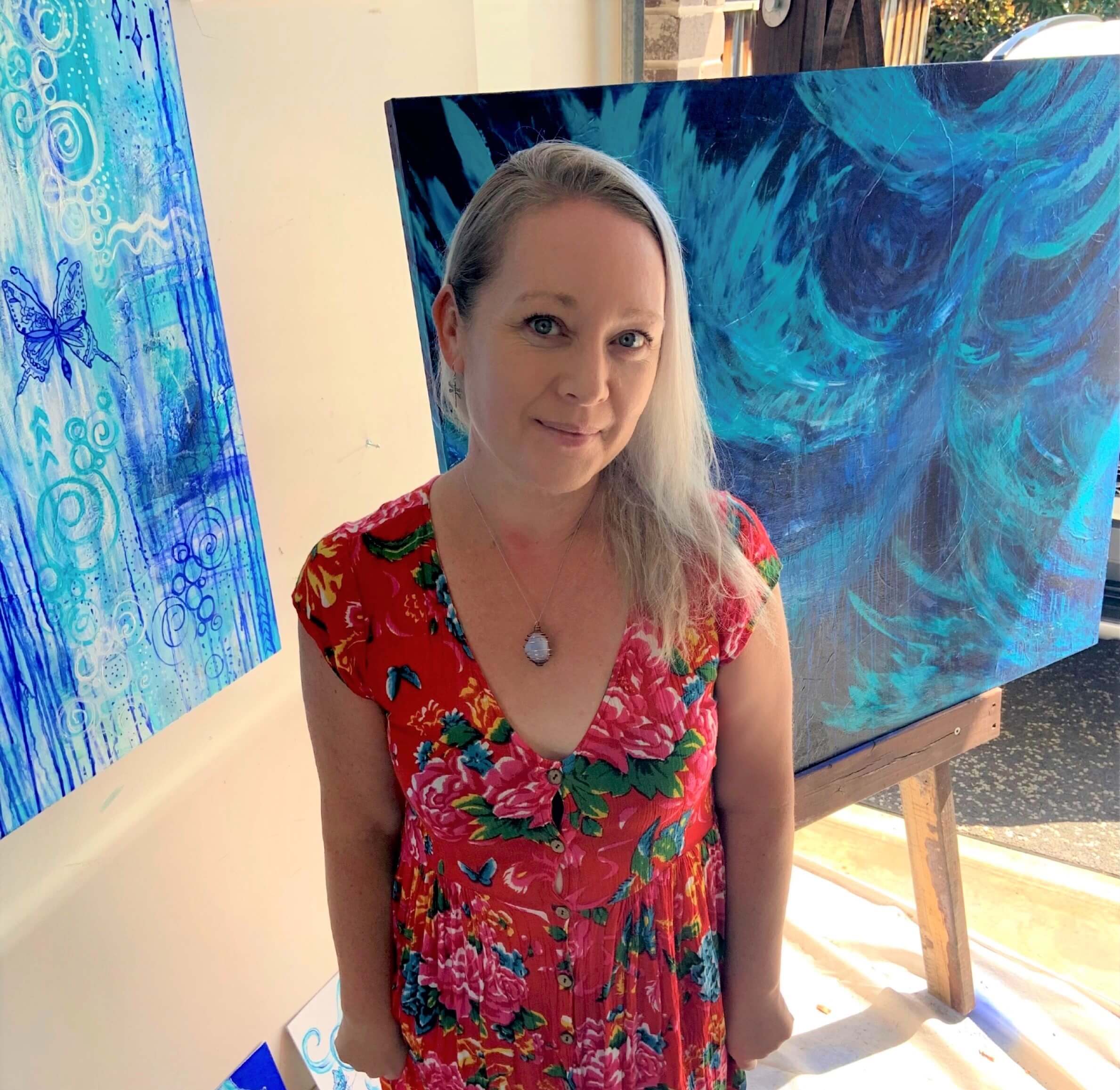 Artist Pauline Nicola standing in her studio with a blue artwork painted on an easel stands behind her.