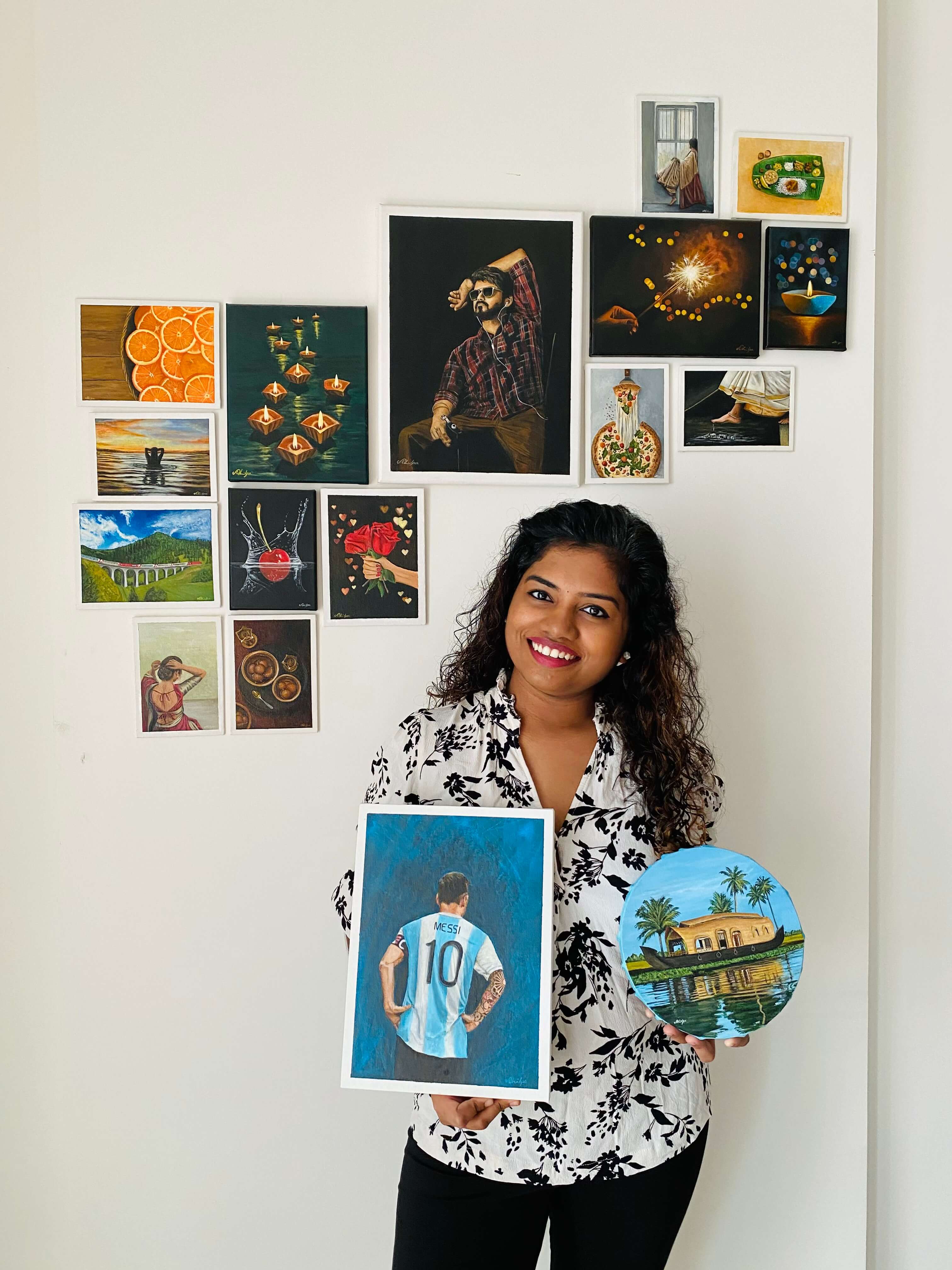 1. Artist Shilpa Rajan smiling and holding two realistic artworks with art behind her on a wall.