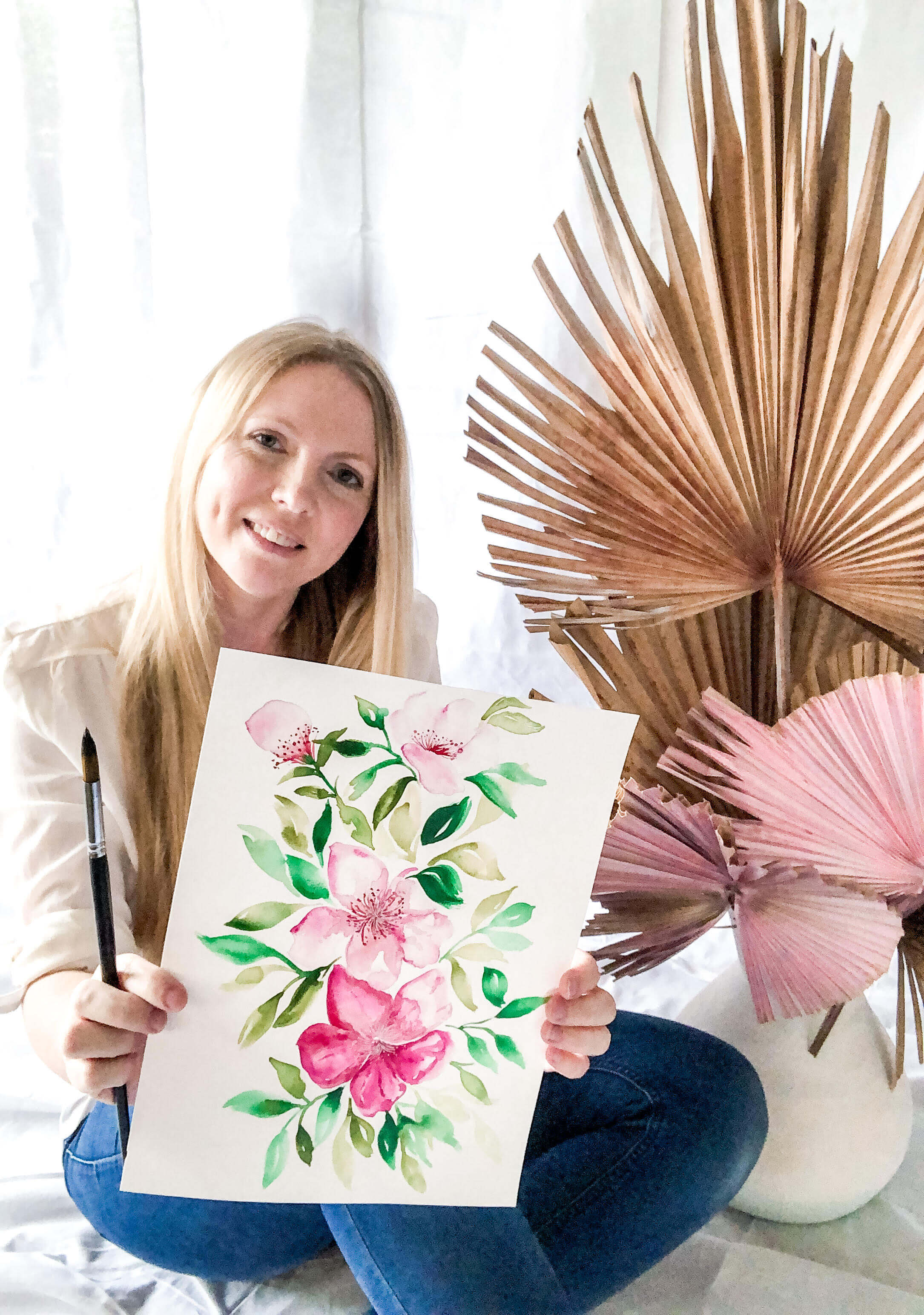 Artist Stacey Bigg holds watercolour painting and watercolour brush and smiles with a dried palm behind her.