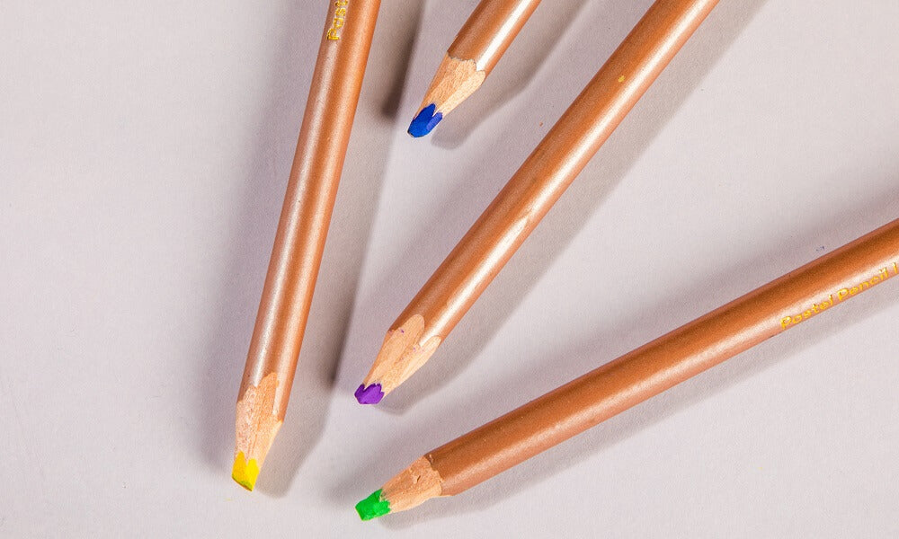 Four pastel pencils of different lengths laying on a white desk. Pencils are in pastel shades of yellow, blue, purple and green.
