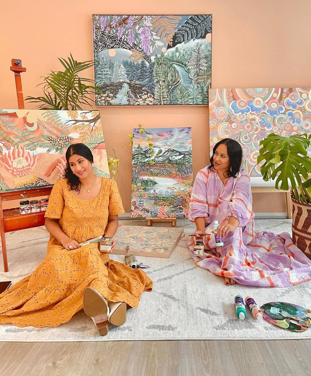 Lani and Zara sitting on the ground of their studio with colourful artworks hanging on the wall behind and on an easel with paints behind them.