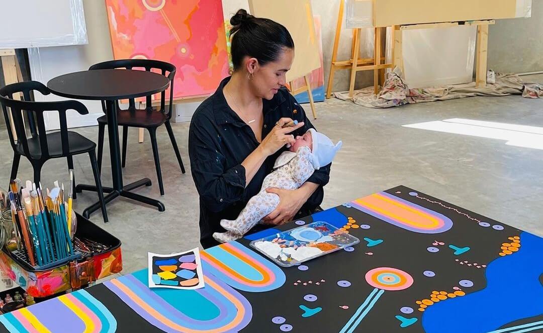 Nardurna holding her baby in an art studio with a coloured artwork in front of her.