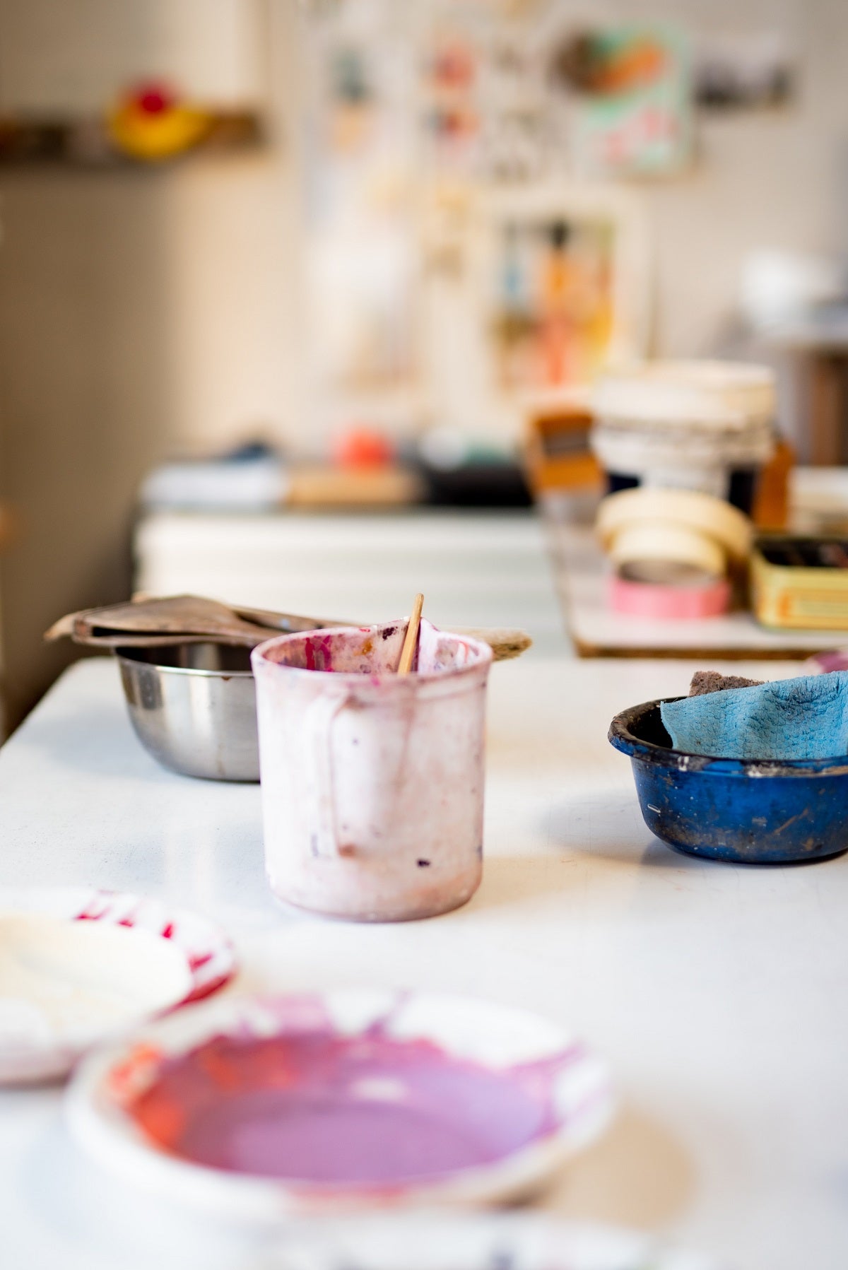 Purple and red acrylic paint in a plastic bowl with two bowls in the background, each with used cloths inside the bowl and a plastic beaker.