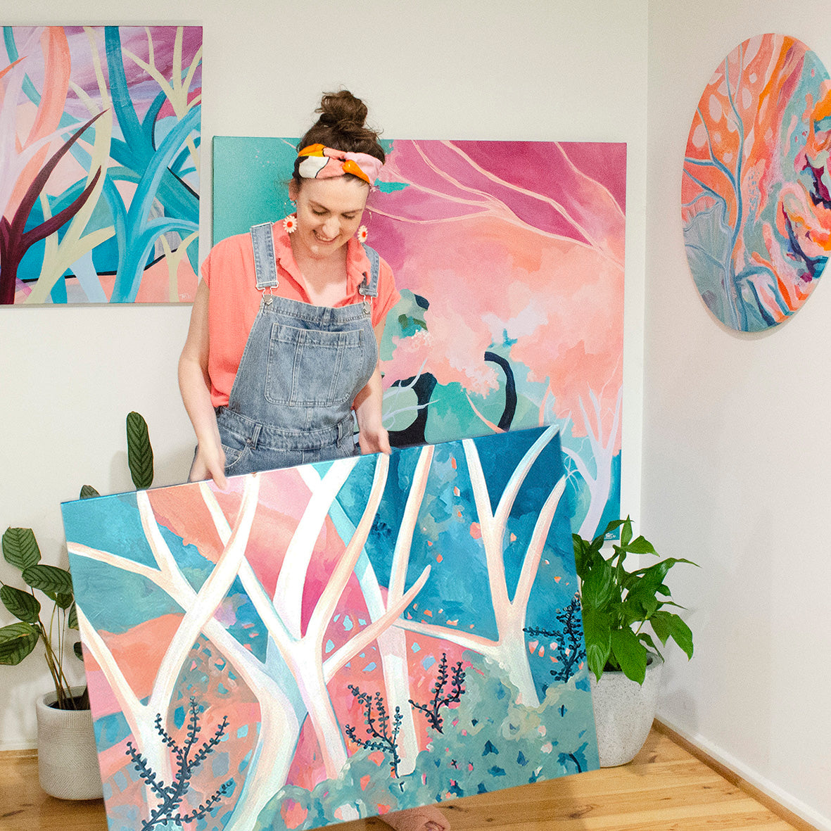 Tessa holding artwork 'a dream I had.'This ia pastel coloured abstract tree painting in her home, Tessa's wearing big flower earrings and a colourful headband.