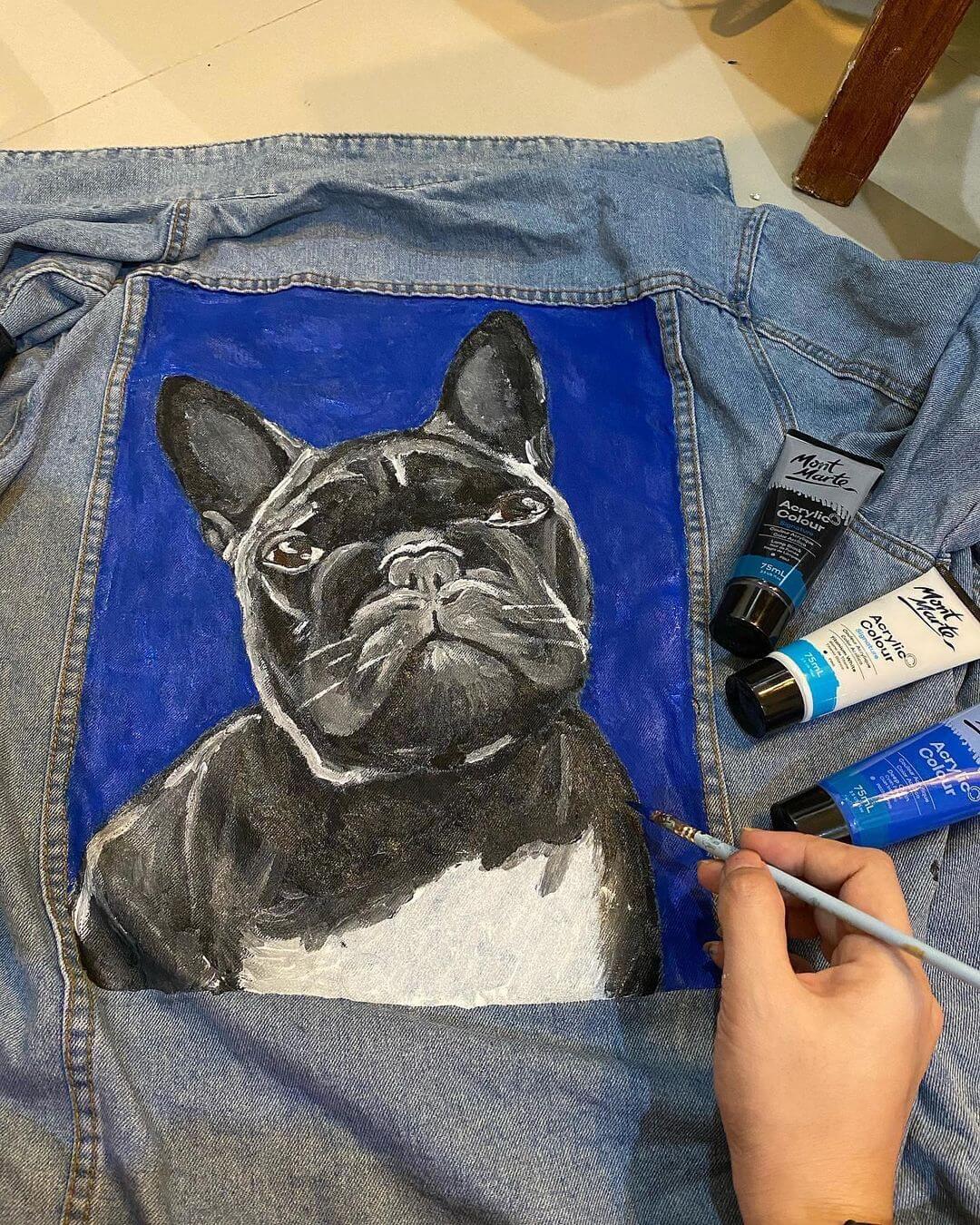 French bull dog painted on the back panel of a blue denim jacket with blue, white and black acrylic paint side by.
