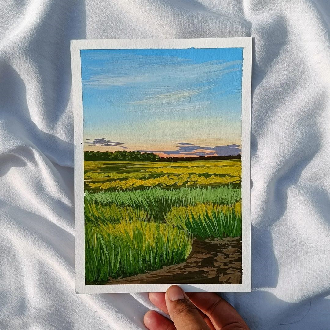 Hand holding a rural landscape of deep green grass and soft blue sky, with white background.