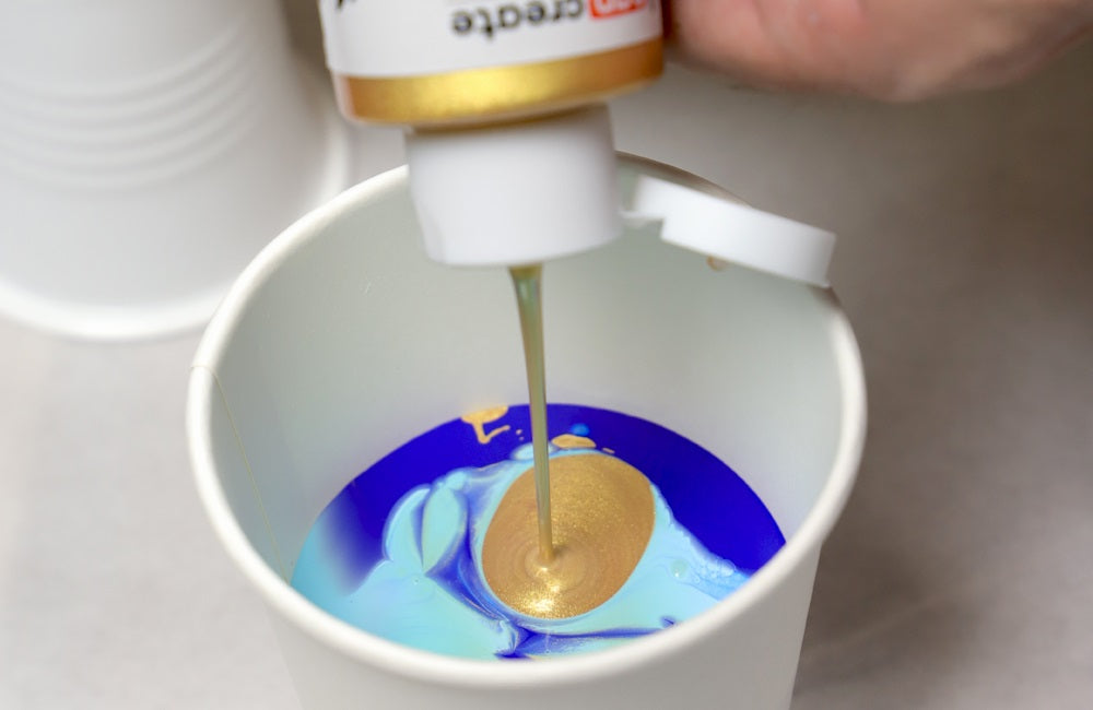 Shades of light and dark blue and gold pour paint being poured into a white plastic cup.