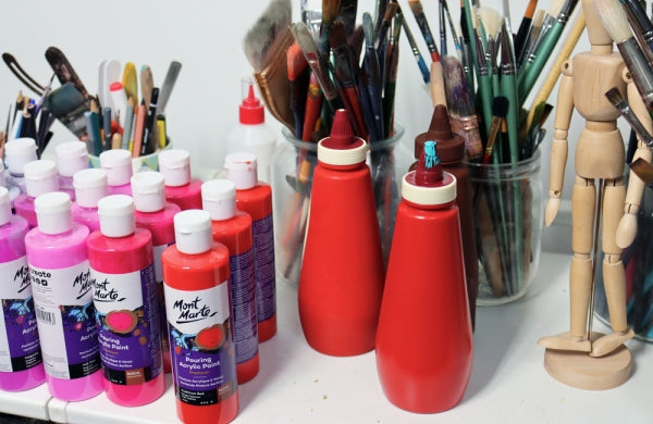 Pink and red bottles of paint, jars of paint brushes and tools, bottles lined up on a table.