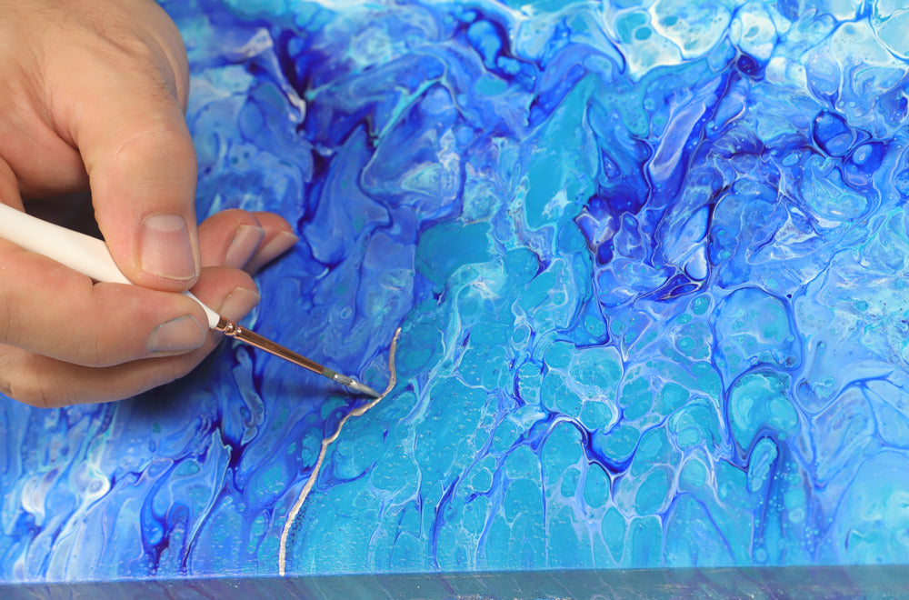 Hand adding foil paint with fine paint brush to painting done in pouring paint.