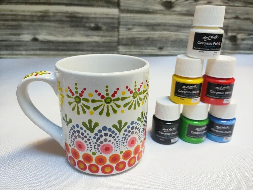 11. A mug painted with a mandela style pattern next to six Mont Marte ceramic paints