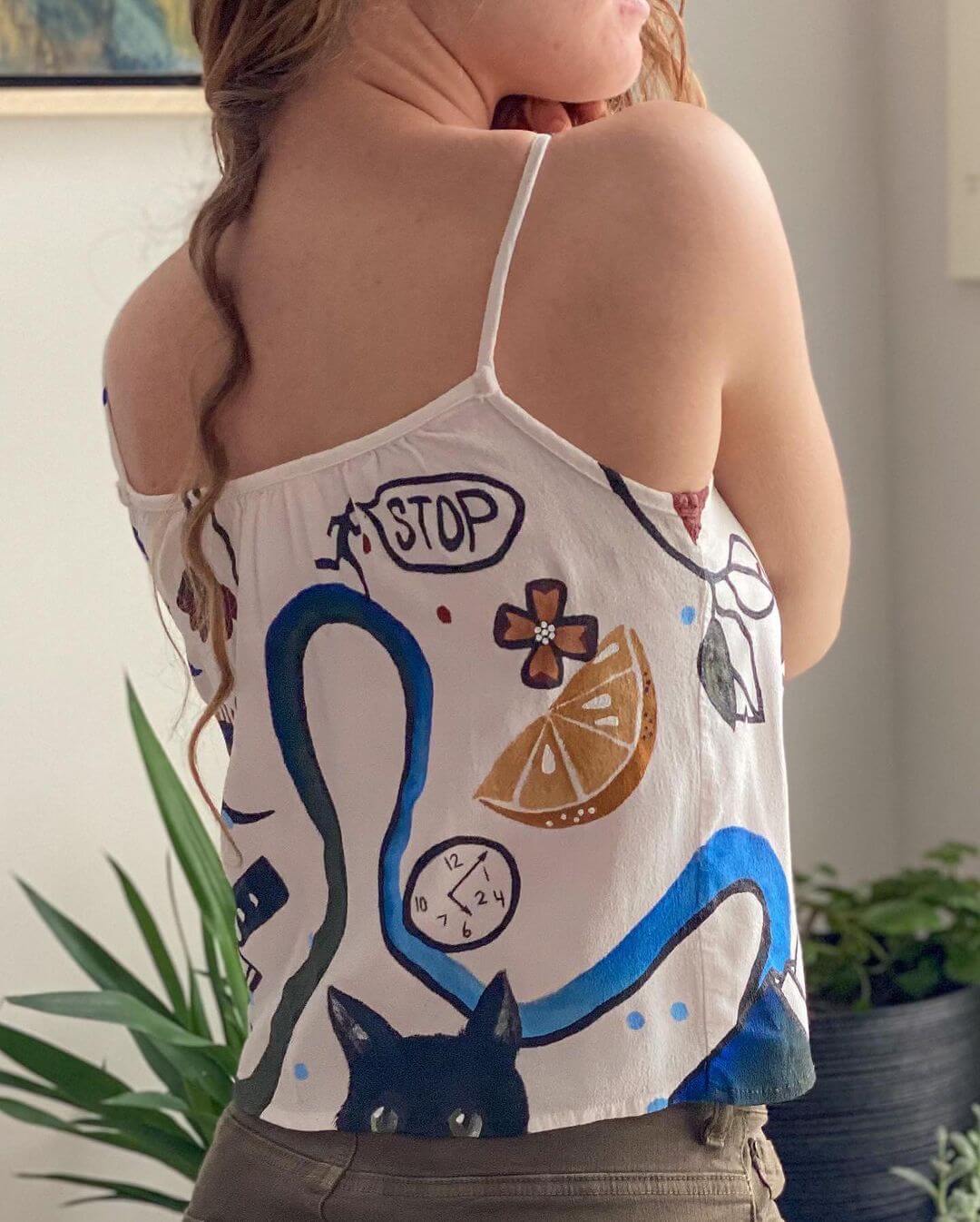 Back of a white singlet with abstract motifs painted throughout of a clock, wave, fruit and stick figure.