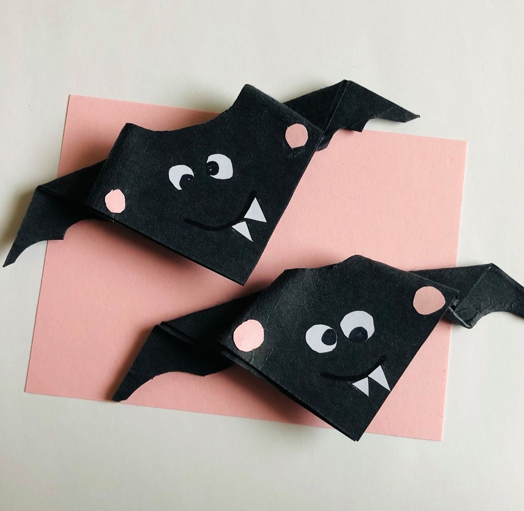 Two black folded origami bats sitting on pink paper.