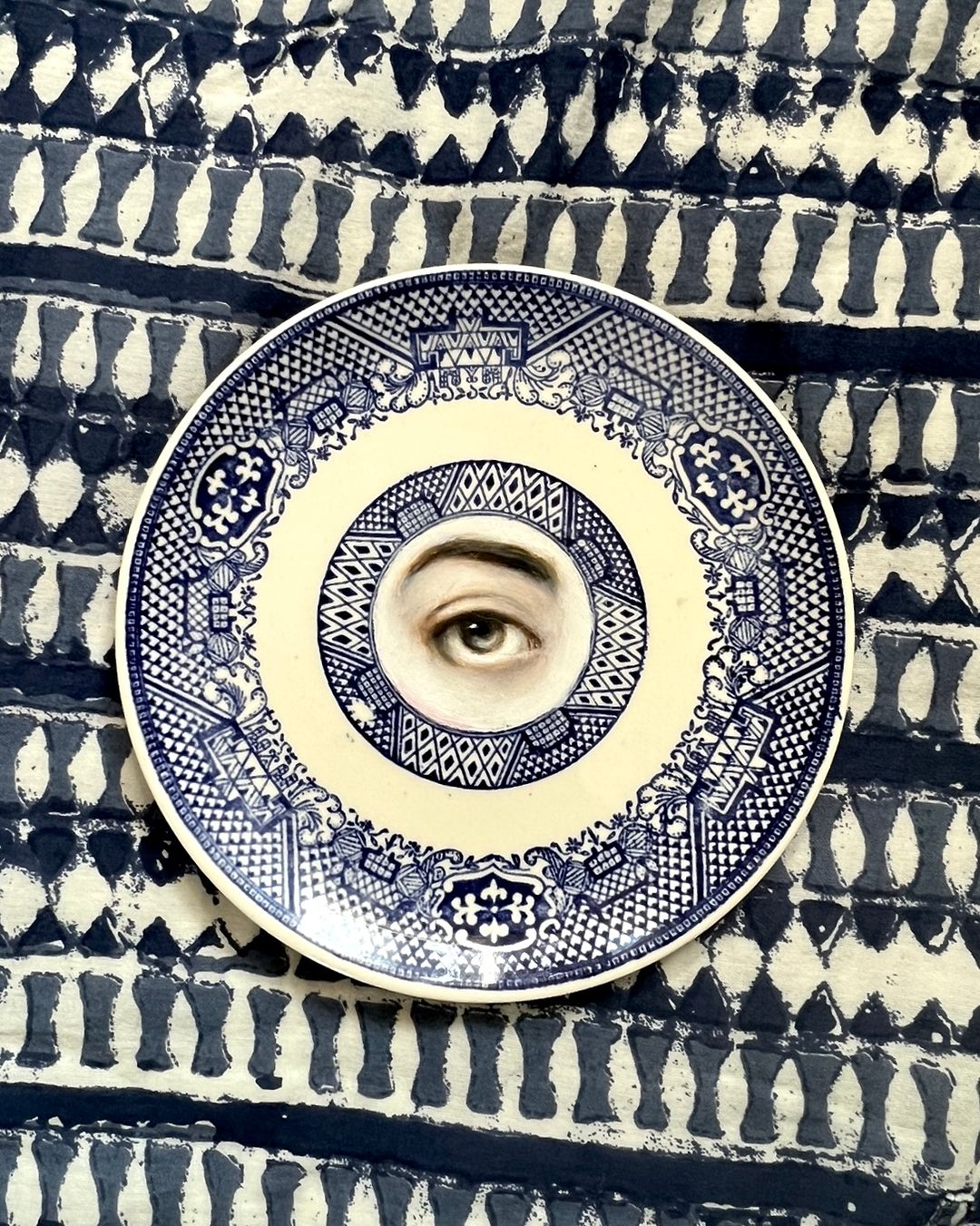 11. @susannah.carson artwork of an eye with an eyebrow in muted colours painted in the centre of a china plate with blue and white designs