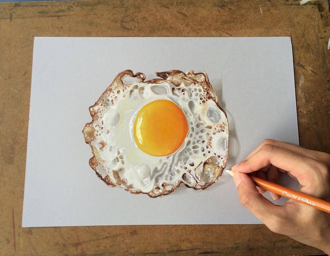 Hand holding a coloured pencil and colouring in a realistic drawing of a fried egg.