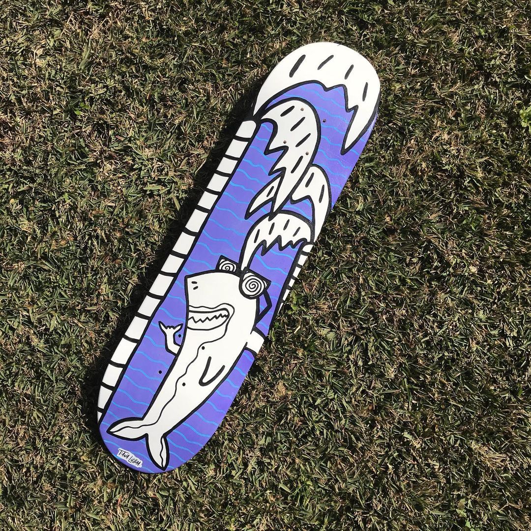 A blue painted skateboard of a shark laying on green grass.