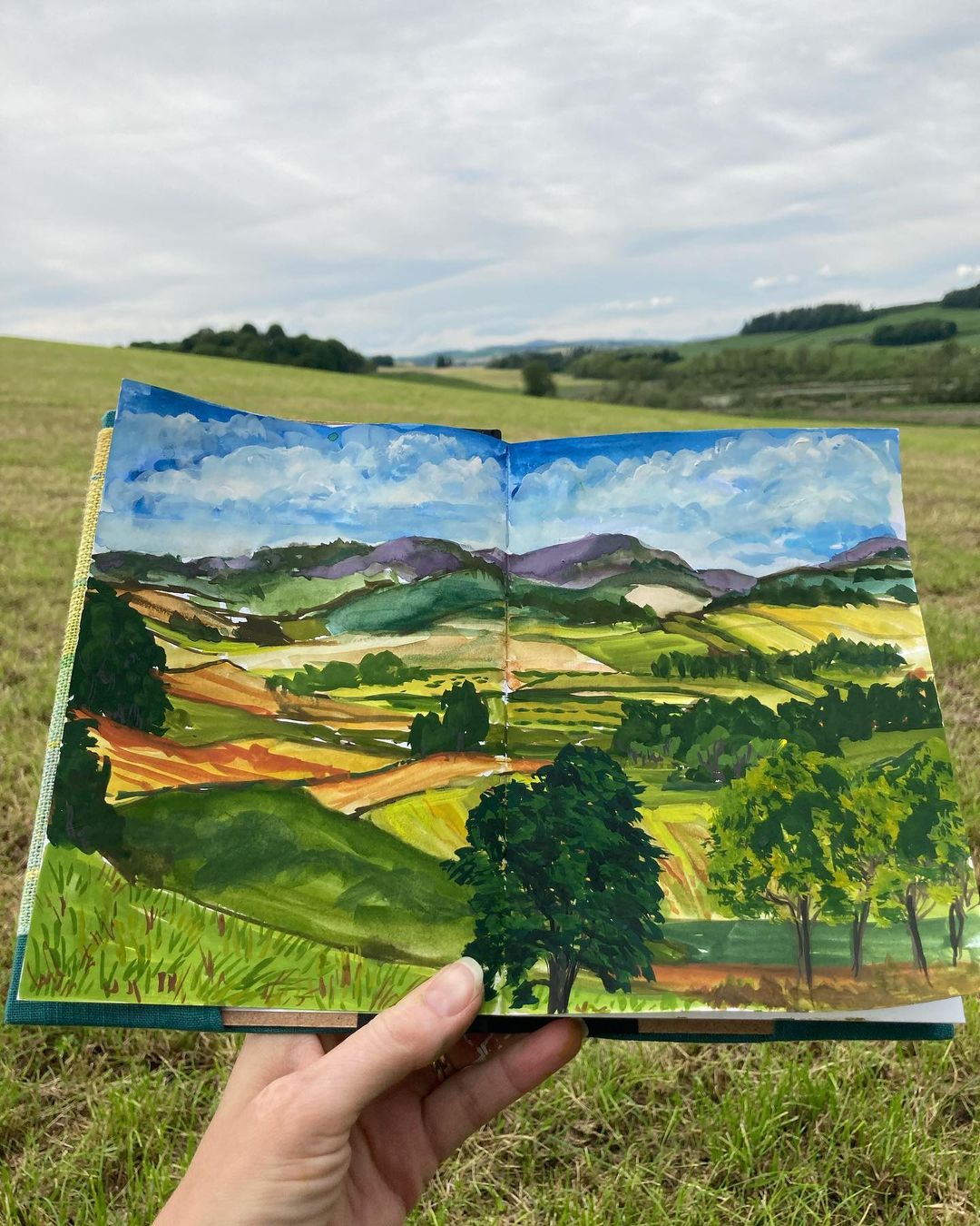 Hand holding a sketchbook of a green rural landscape painted in gouache.