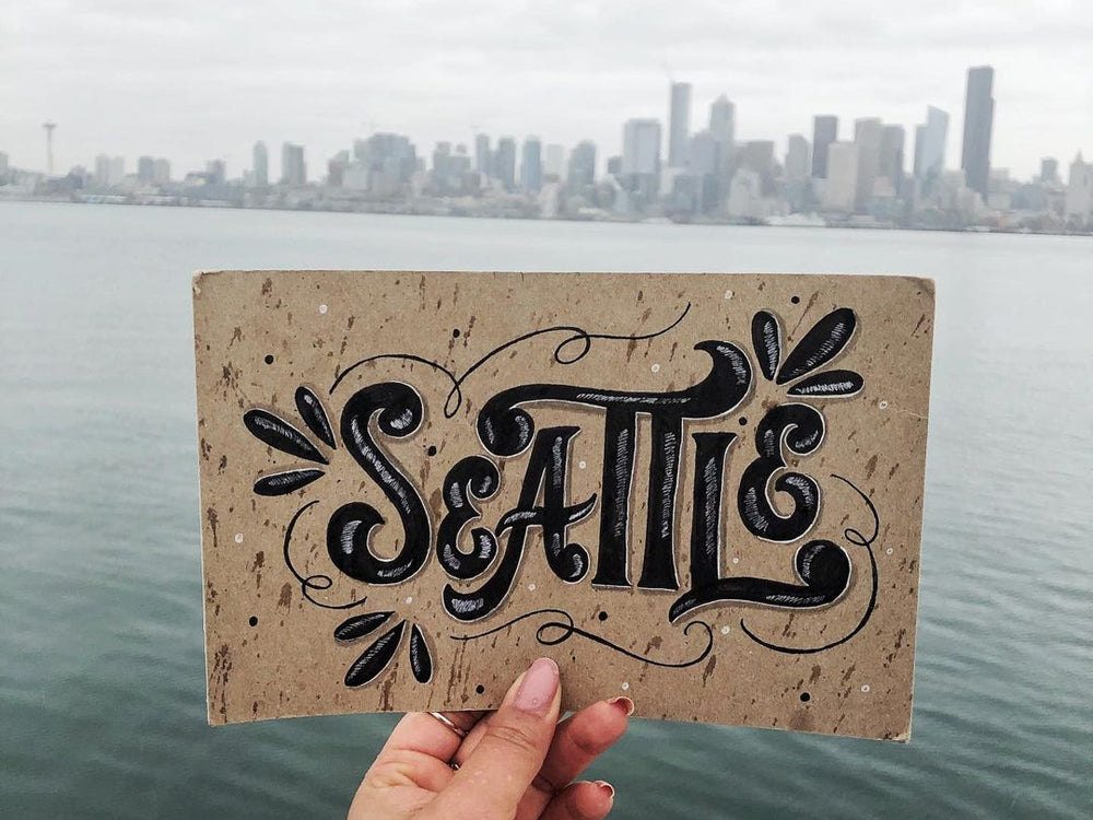 Hand holding a sign with "Seattle" on it, with the cityscape in the background.