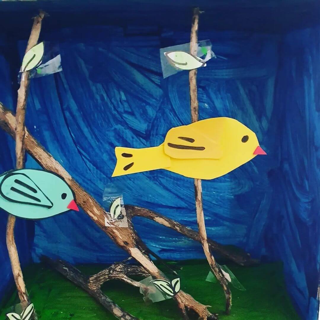 A yellow and blue fish made from paper and stuck on sticks in a shoe box to look like a mini aquarium.