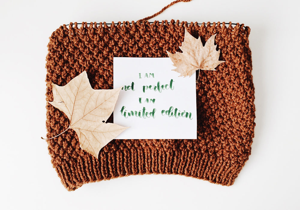 Green hand lettering on a piece of white paper, lying on a knitted beanie with maple leaves on it.