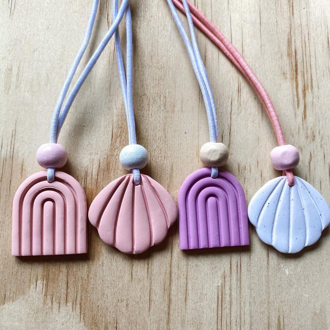 Four clay necklaces with a dusty pink rainbow and shell, purple rainbow and light blue shell. Each with a bead on blue and red string.