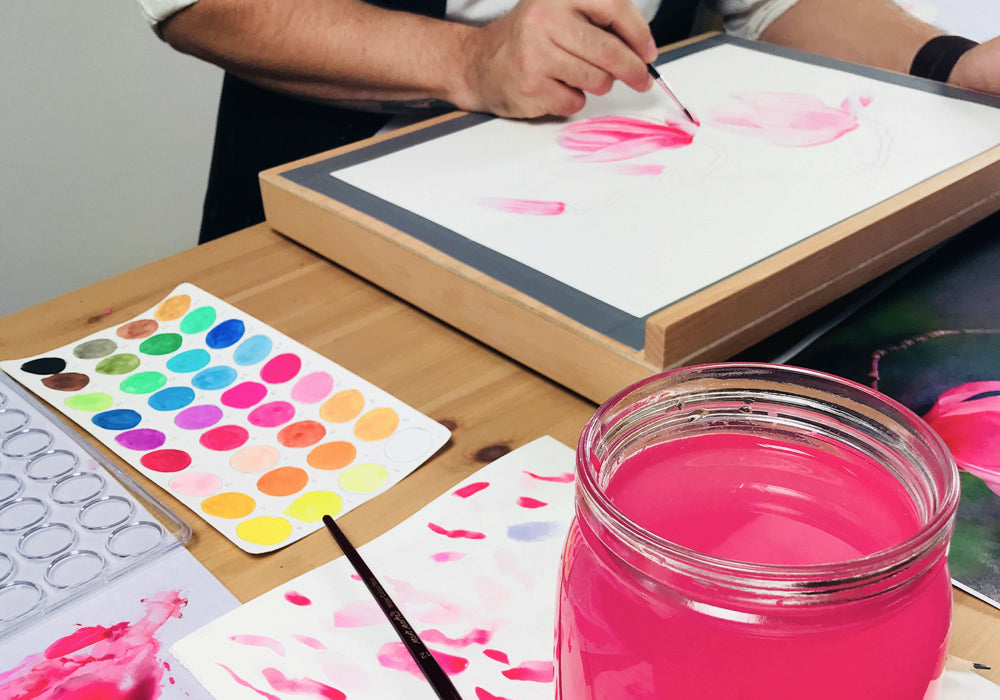 Man painting pink water colour flower on one half of desk, watercolour chart and pink jar of water is on the other half of the desk.