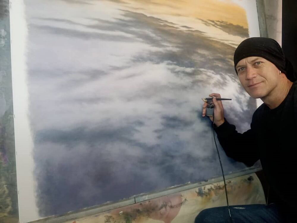 Dawie Mocke smiling at camera while spraying clouds on a landscape artwork.