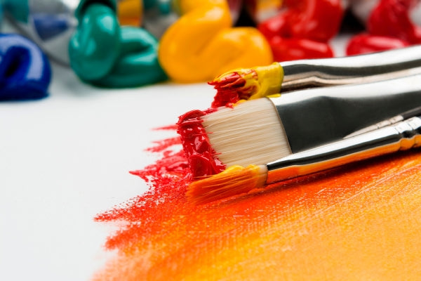 Paint brushes with red and orange paint still on them lying on a page partially painted in red, orange and yellow near blobs of green, blue, yellow and red paint.