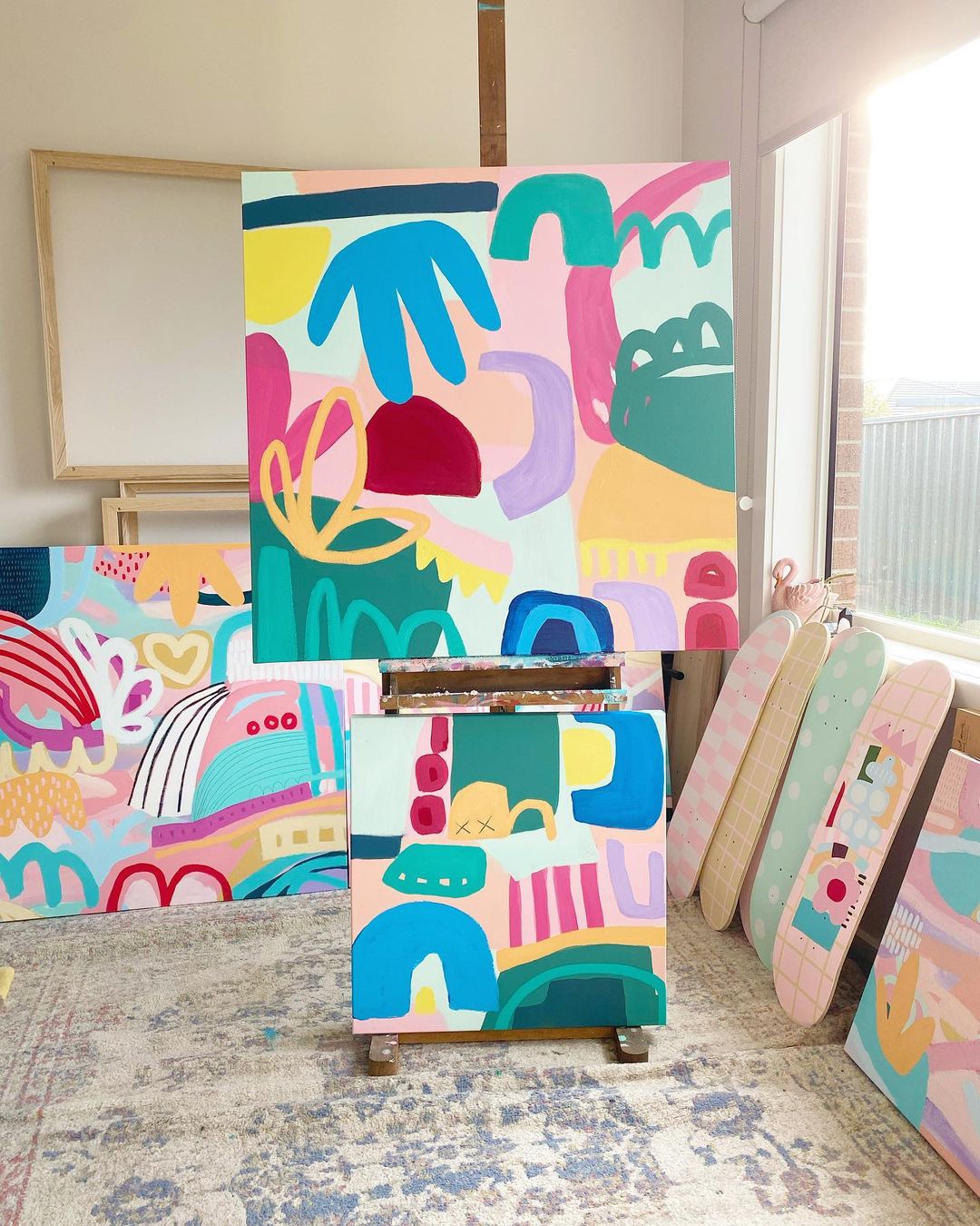 An abstract pastel artwork on an easel with skateboards next to the easel on the wall all painted with a pastel abstract design.