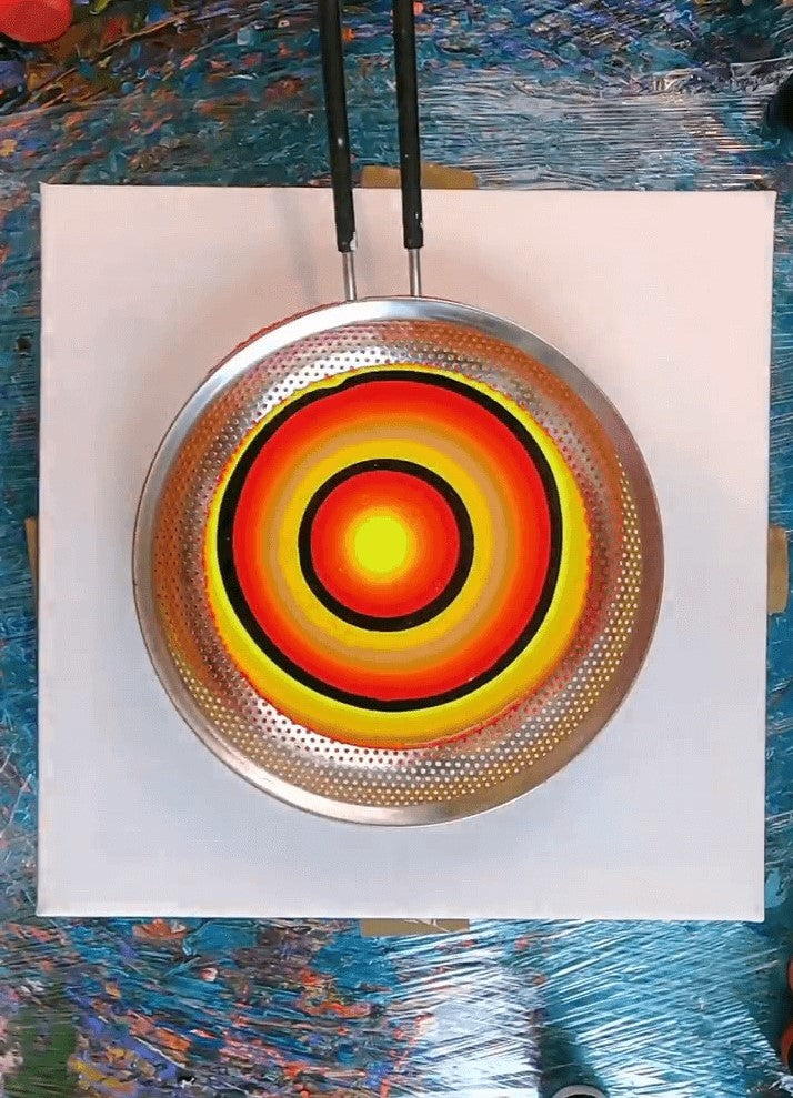 2. A strainer on a white canvas with circles of red, yellow, gold and black paint sitting on the strainer.