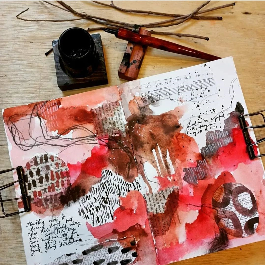 An art journal with layers of pink and red abstract paint with a calligraphy pen and a branch near by.