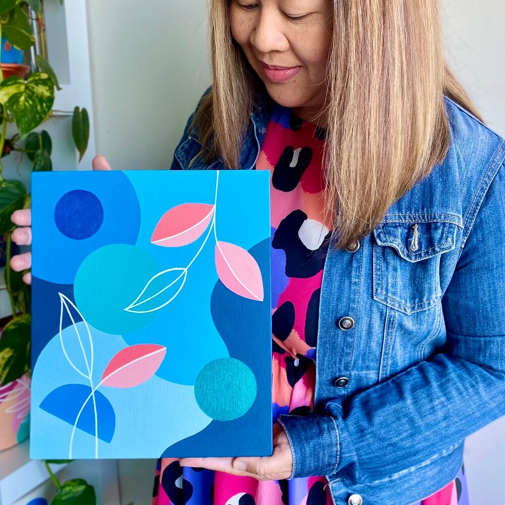 Ann in a blue denim jacket holding a blue artwork with pink leaves painted on it.