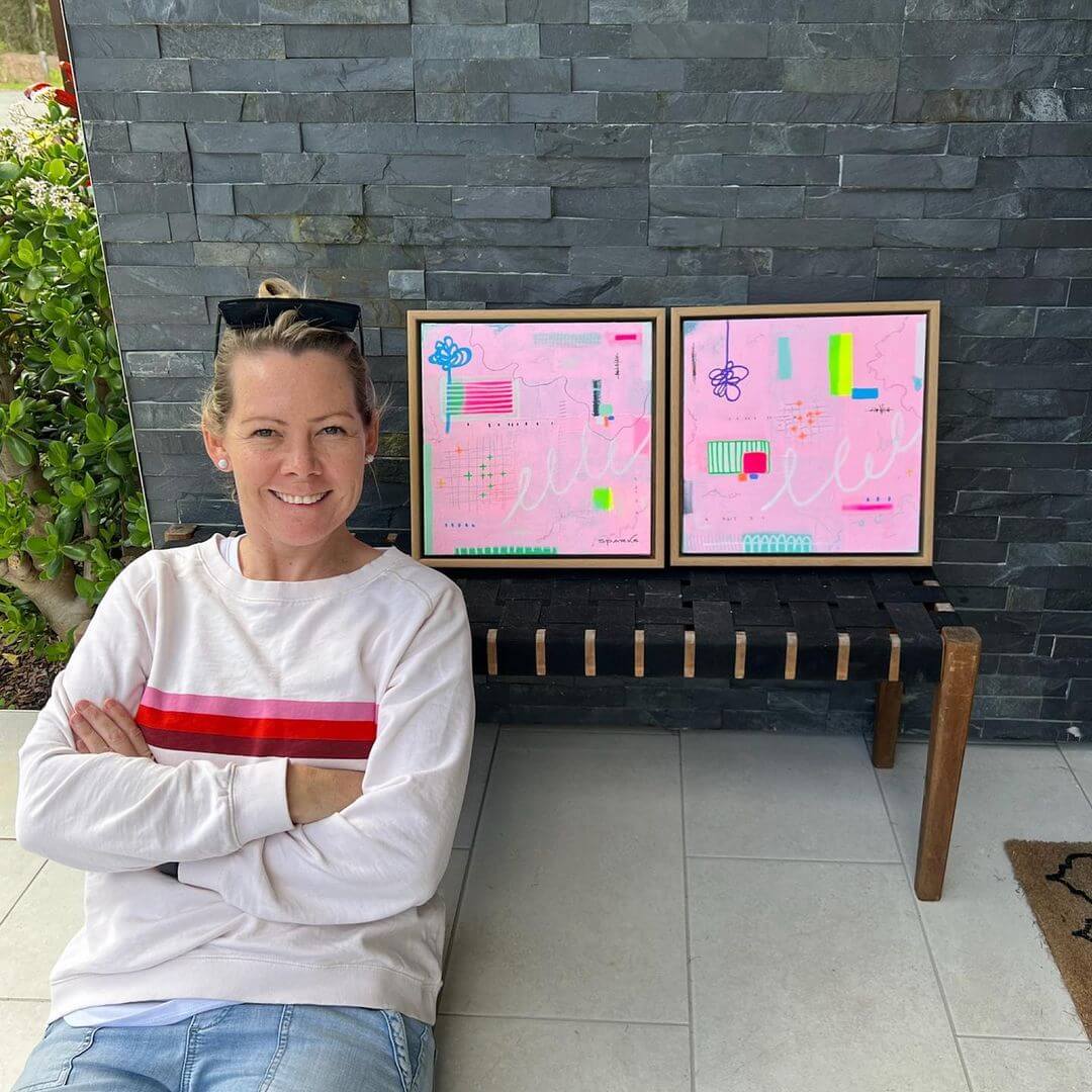 Artist Michelle Sparks smiling next to two of her framed artworks.