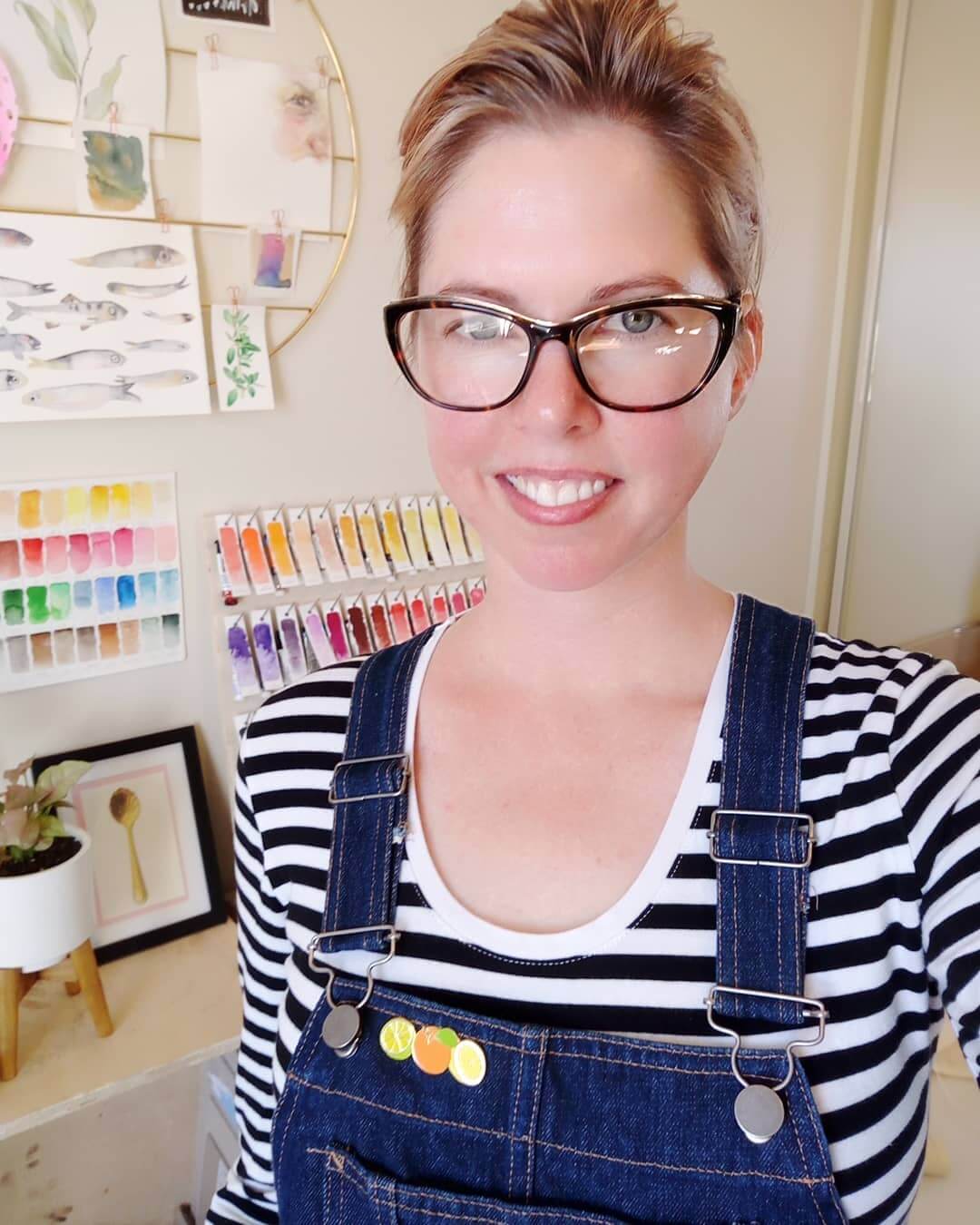 Artist Sara Brenton smilling in her studio with an apron on and various watercolour swatches behind her.