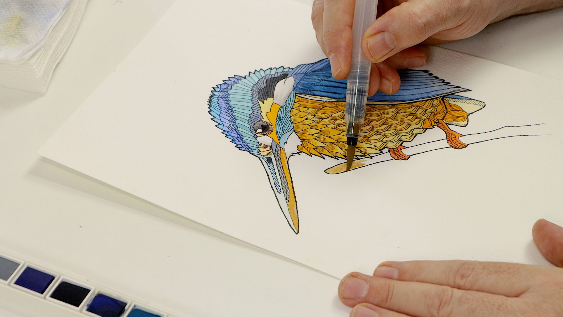 Hand holding a watercolour brush painting a stylised kingfisher artwork