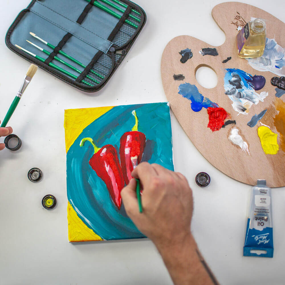 Hand painting an oil paint artwork of two colourful chillis with a wooden palette and brushes next to it.
