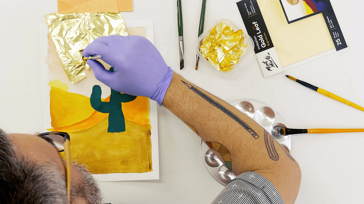 2. Man painting gold leaf onto an existing gouache artwork of a cactus in the desert.