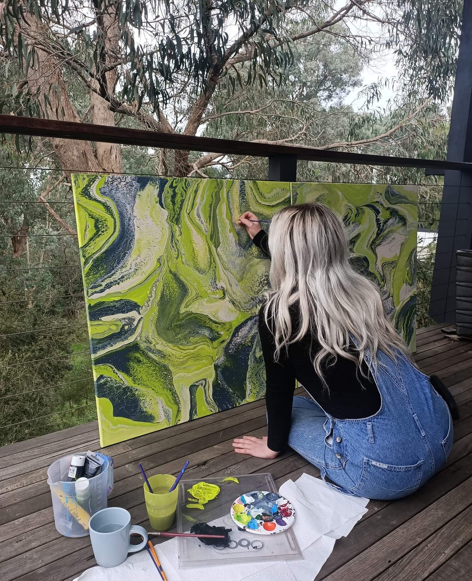 Steff's painting a green artwork called 'Northern lights.' Steff's sitting on wooden floor boards wearing denim overalls and painting with a brush.