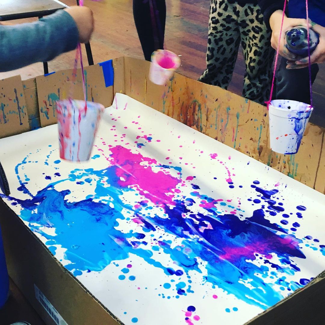 2. @lilcraftergroup pendulum swing painting with pink and blue paints spilling from cups on a white surface