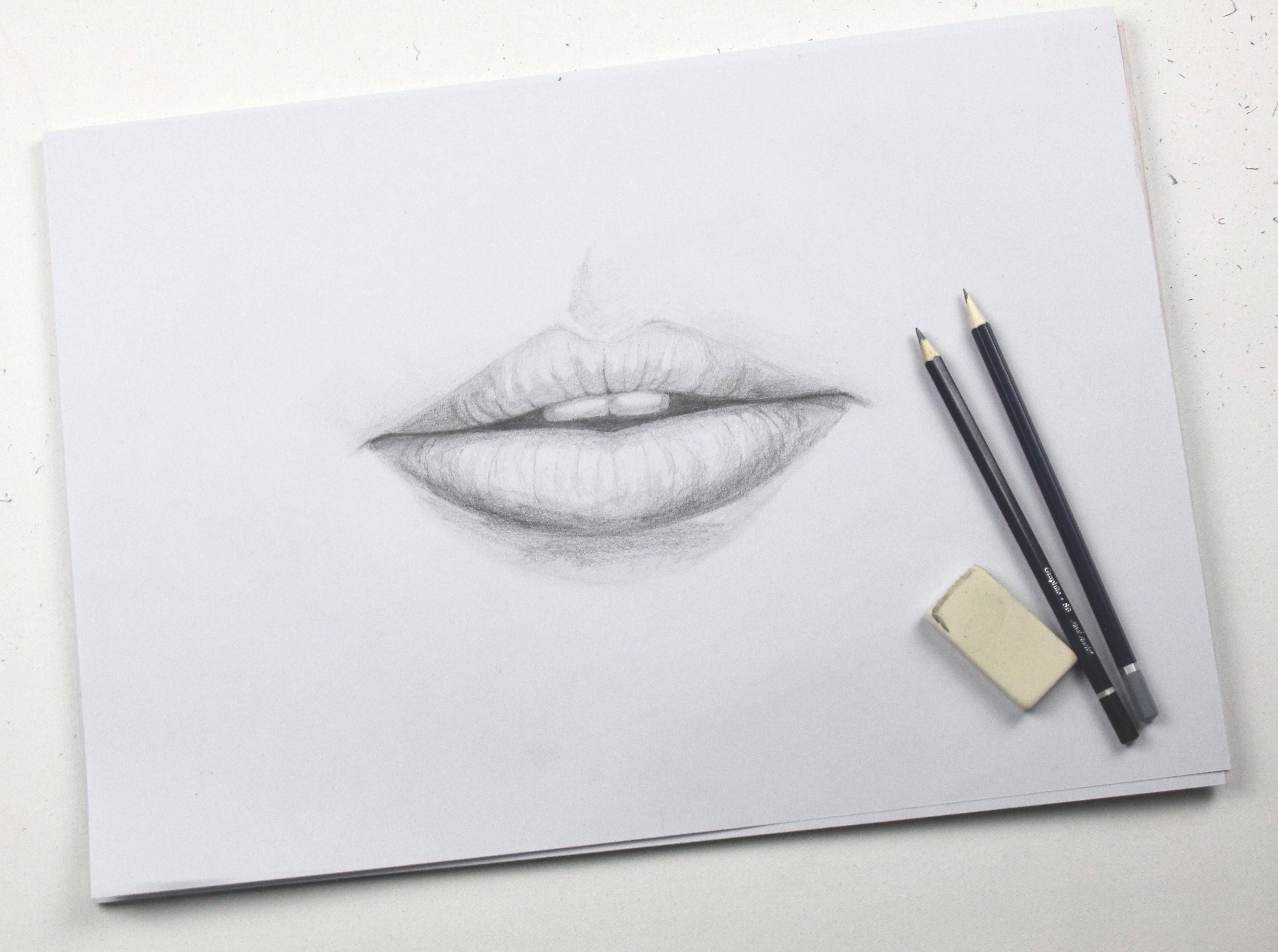 Drawing of lips drawn in graphite on white paper next to two pencils and an eraser.