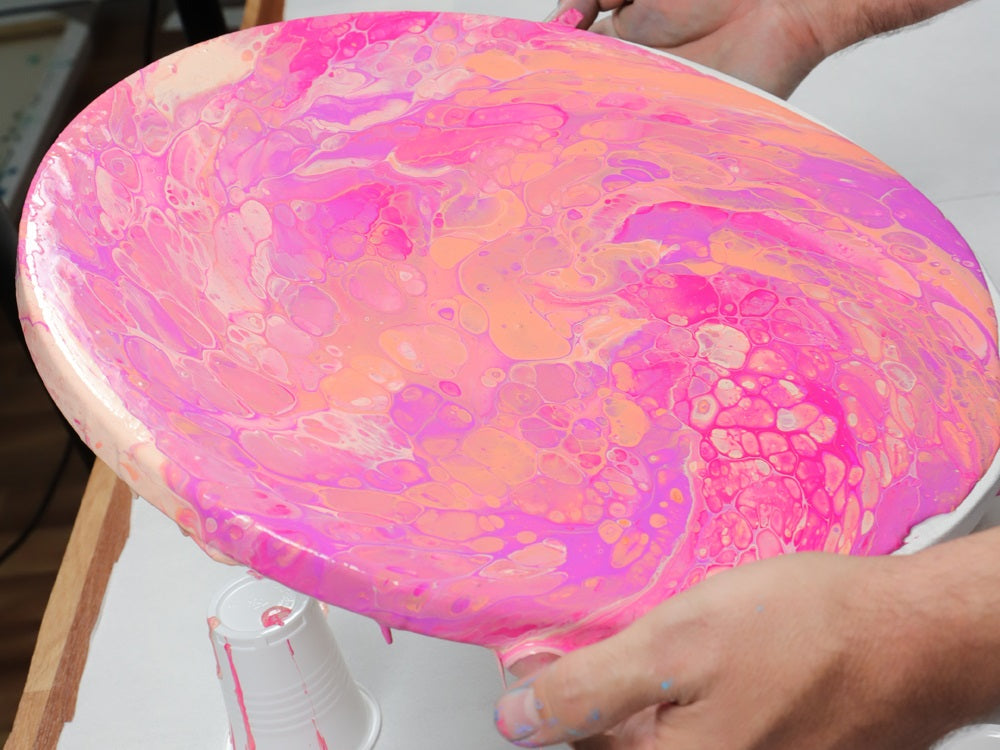 A hand holding a pink marbled pour painting on a circular canvas.