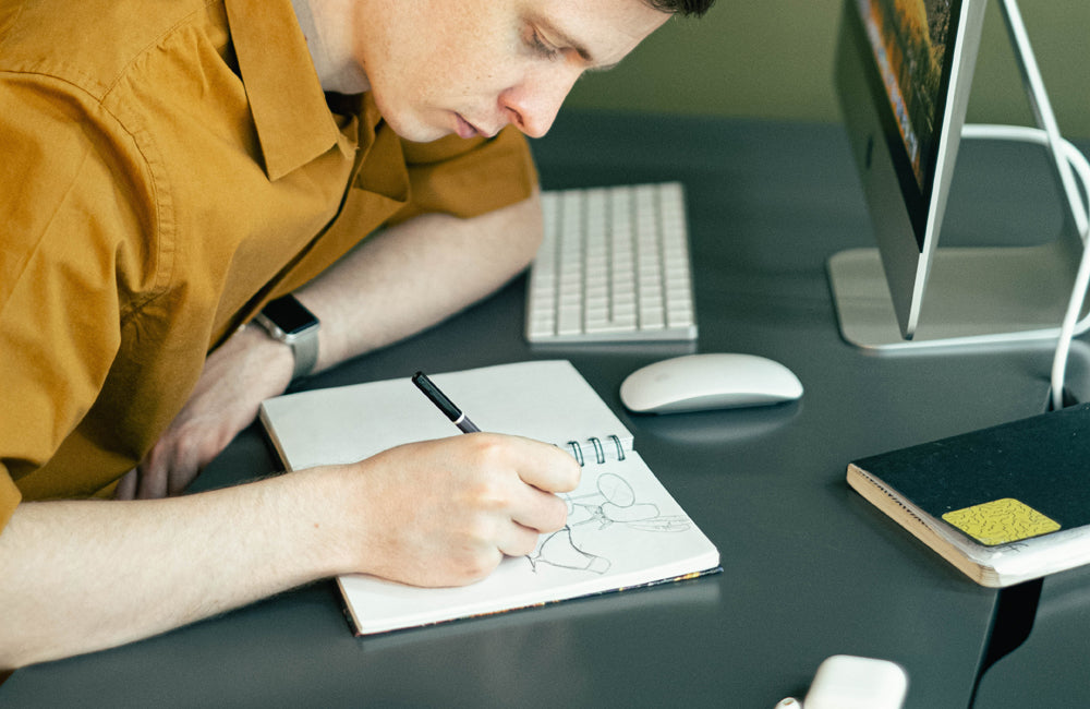 Male wearing a yellow shirt, sketching in a notebook next to a computer.