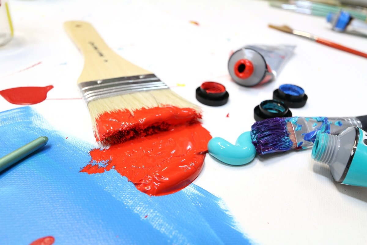 A paint brush in red paint with various blue paints spread out on a white surface.