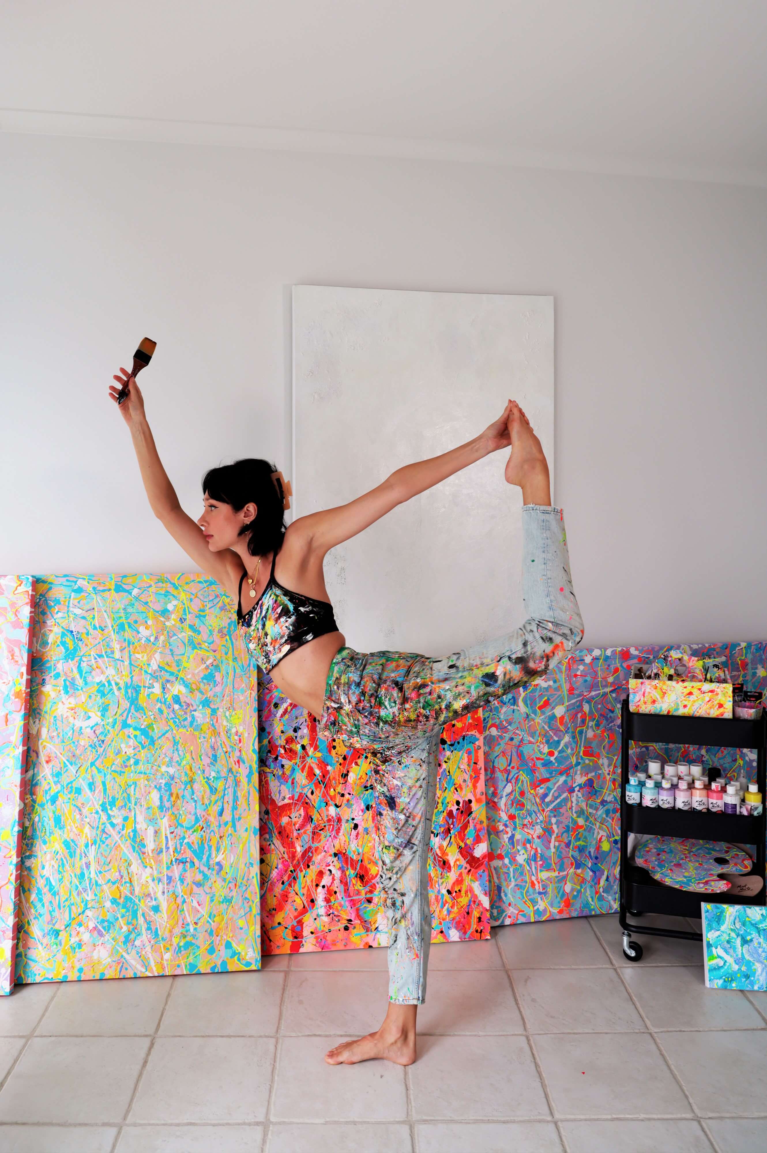 Bridget holding a paint brush in one hand and doing a yoga pose with the other. In the back are her artworks and an art trolley